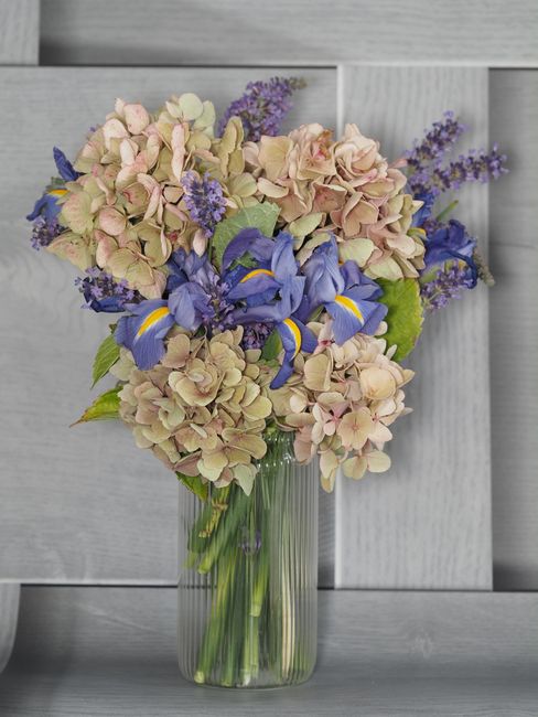 A bouquet of soft pink and cream hydrangeas, accented with vivid purple irises and sprigs of lavender flowers, arranged in a clear glass vase. The bouquet shows lush, fresh petals and green leafy stem