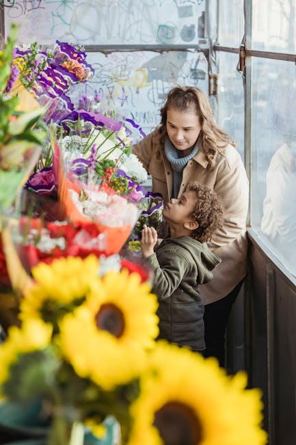 A woman and a young girl are inside a flower shop, surrounded by a vibrant display of fresh flowers. The woman has wavy light brown hair and is wearing a beige coat over a blue turtleneck, while the g