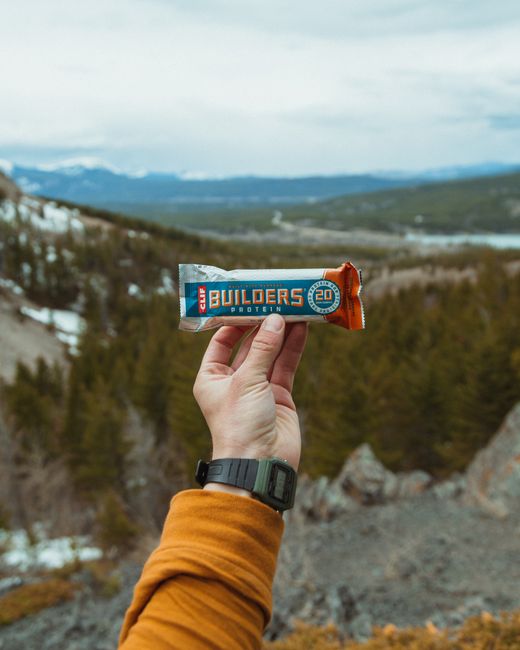 A person's hand holding a protein bar labeled 'Builders' with the number 20, positioned in front of an expansive outdoor landscape featuring rolling hills, scattered trees, and distant mountains under