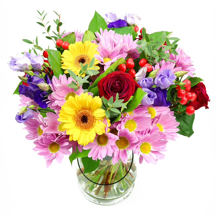 This image shows a generously sized, mixed-colour bouquet arranged in a clear glass vase, set against a pristine white background that draws attention to every detail. At its centre, several deep red roses bloom in velvety spirals, bringing a sense of classic romance and depth, ideal for expressing affection to someone special in Teddington. Beside them, two bright yellow gerbera daisies with warm, brown-orange centres stand out like little suns, lending the piece a cheerful, optimistic character that recalls the golden light over Bushy Park in late afternoon. Soft pink daisies or chrysanthemums encircle these focal blooms, their many fine petals and small yellow centres forming a cushion of pastel colour that softens and balances the stronger tones. Woven throughout the arrangement, lisianthus blossoms in shades of purple and lavender introduce graceful, bell-shaped flowers with a slightly cooler palette, adding sophistication and visual depth. Between petals and leaves, clusters of glossy red hypericum berries appear like small jewels, offering a rounded texture and a modern twist. The bouquet is framed and supported by a mix of rich green foliage-broad, dark leaves give structure and presence, while lighter, more delicate sprigs and fronds bring movement and a gently untamed, garden-fresh feel, reminiscent of riverside walks along the Thames toward Kingston. The transparent vase reveals an orderly fan of neatly cut stems in clear water, evidence of professional care and freshness from a trusted Teddington florist. The overall atmosphere is one of joyful abundance and thoughtful design, making this bouquet perfectly suited for birthdays, thank-yous, or simply brightening a TW11 home with colour and life.